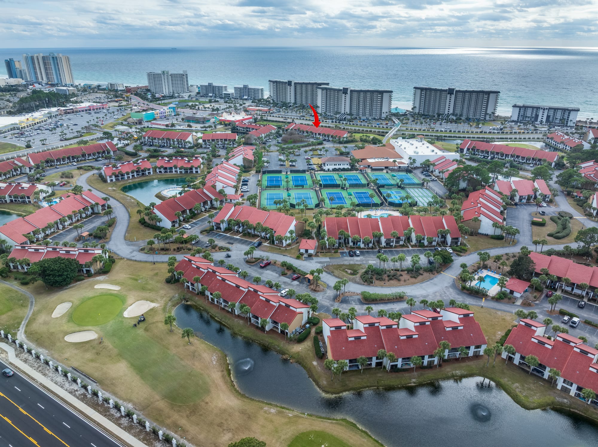 Aerial of red-roof buildings, tennis courts, a pond, and the gulf. A red arrow points toward a group of buildings.
