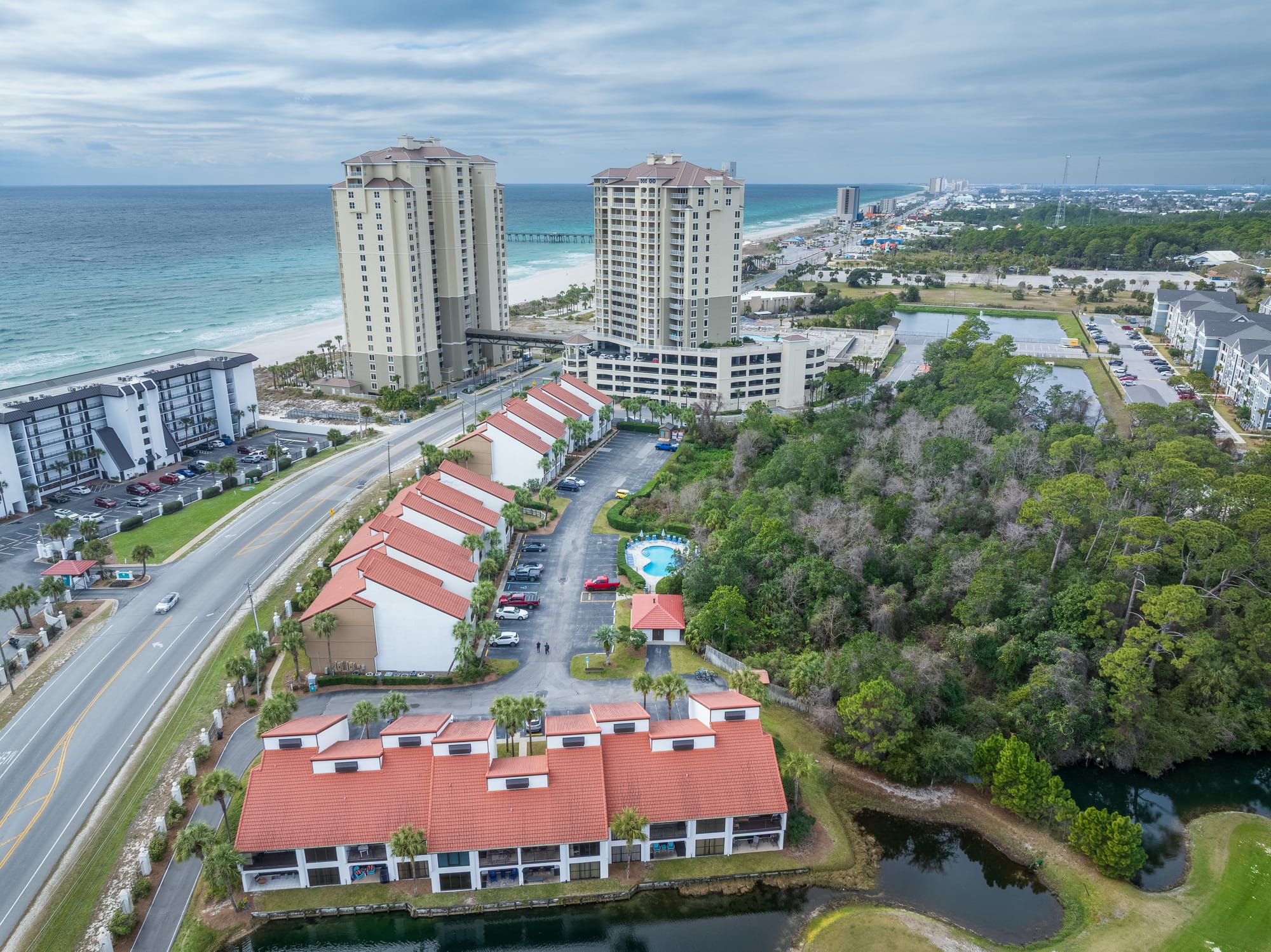 Aerial of red-roof buildings, a small pool, trees, and beachfront towers along the gulf.
