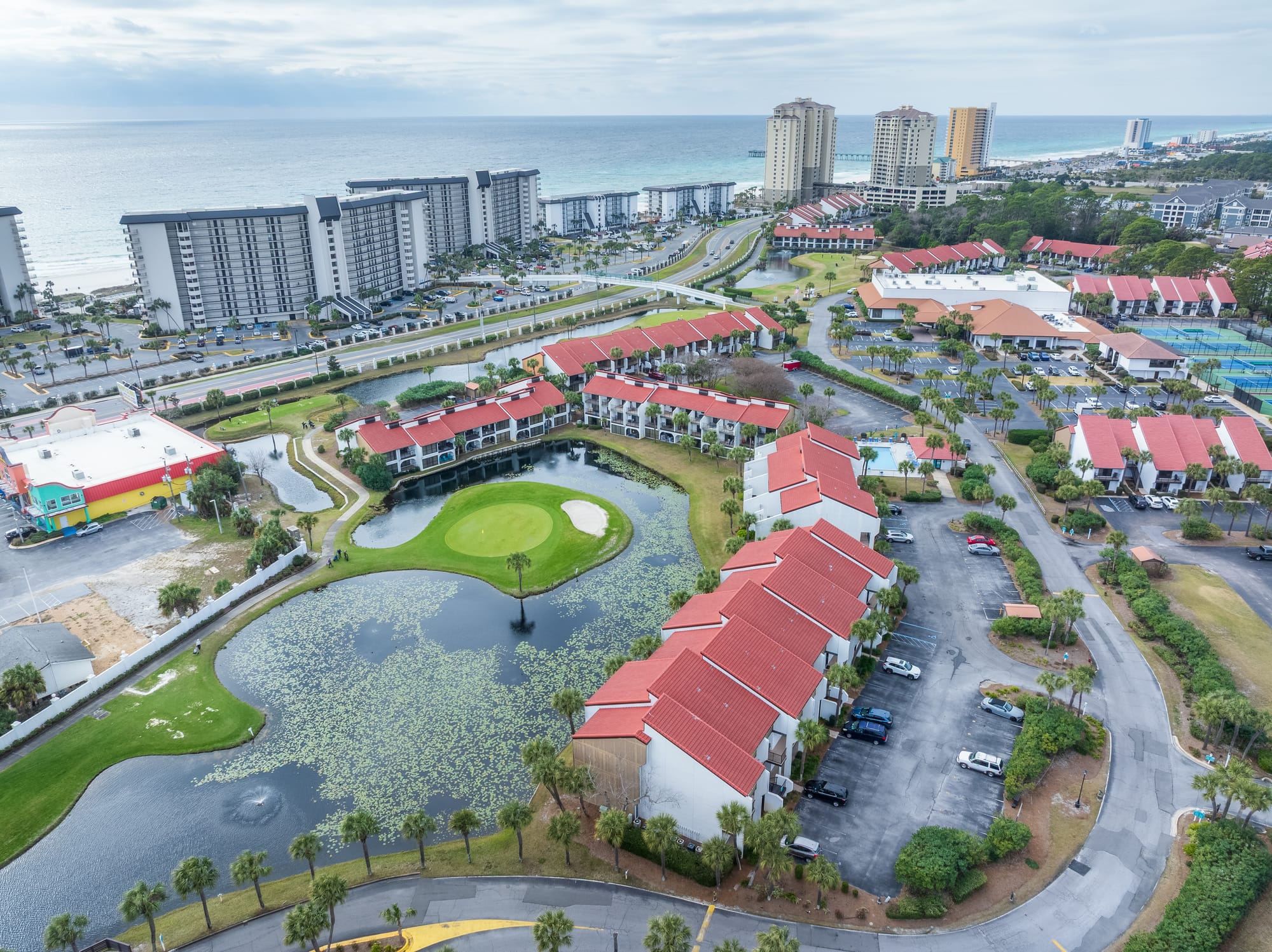 Aerial of a golf green surrounded by water, red-roof buildings, and the gulf in the distance.