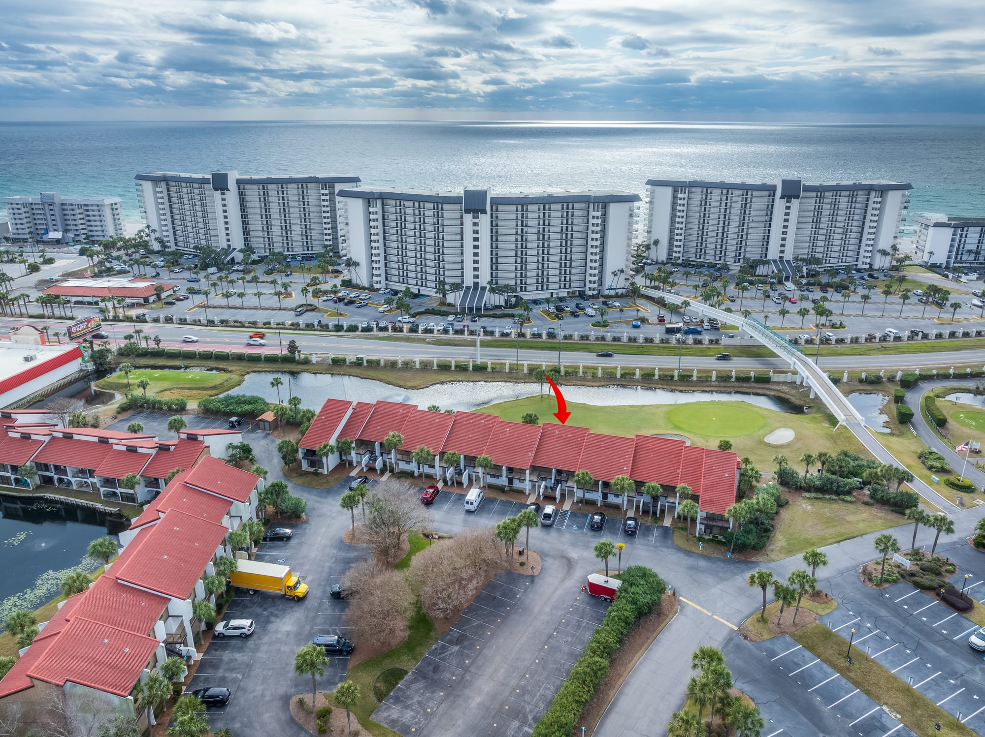Closer aerial of red-roof buildings and the gulf beyond a road. A red arrow points at one roof.
