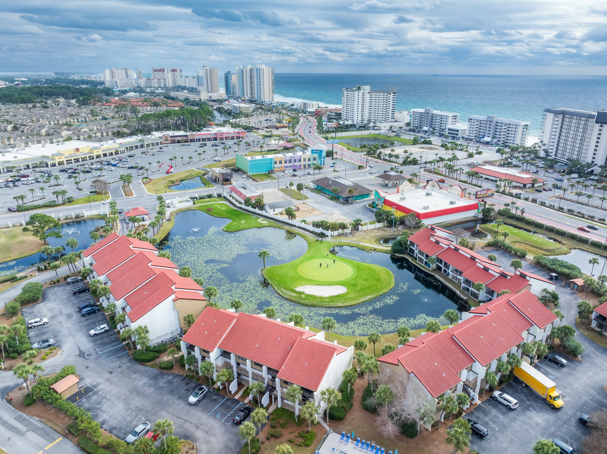 Aerial looking across a pond and golf green toward the beachfront high-rises and the gulf.