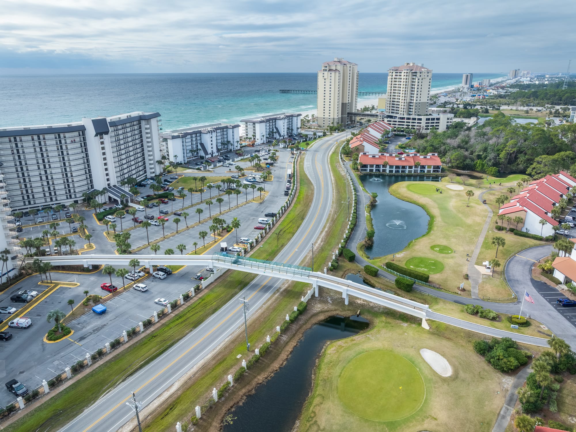 Aerial of a pedestrian bridge crossing a road, with the gulf and beachfront towers beyond.