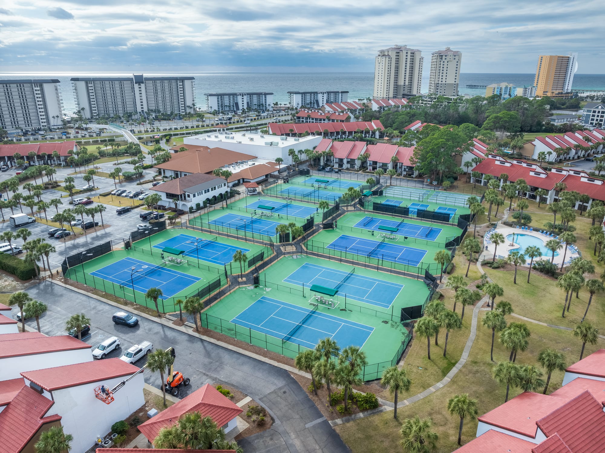 Aerial of several blue and green tennis courts, a pool, palm trees, and the gulf beyond.