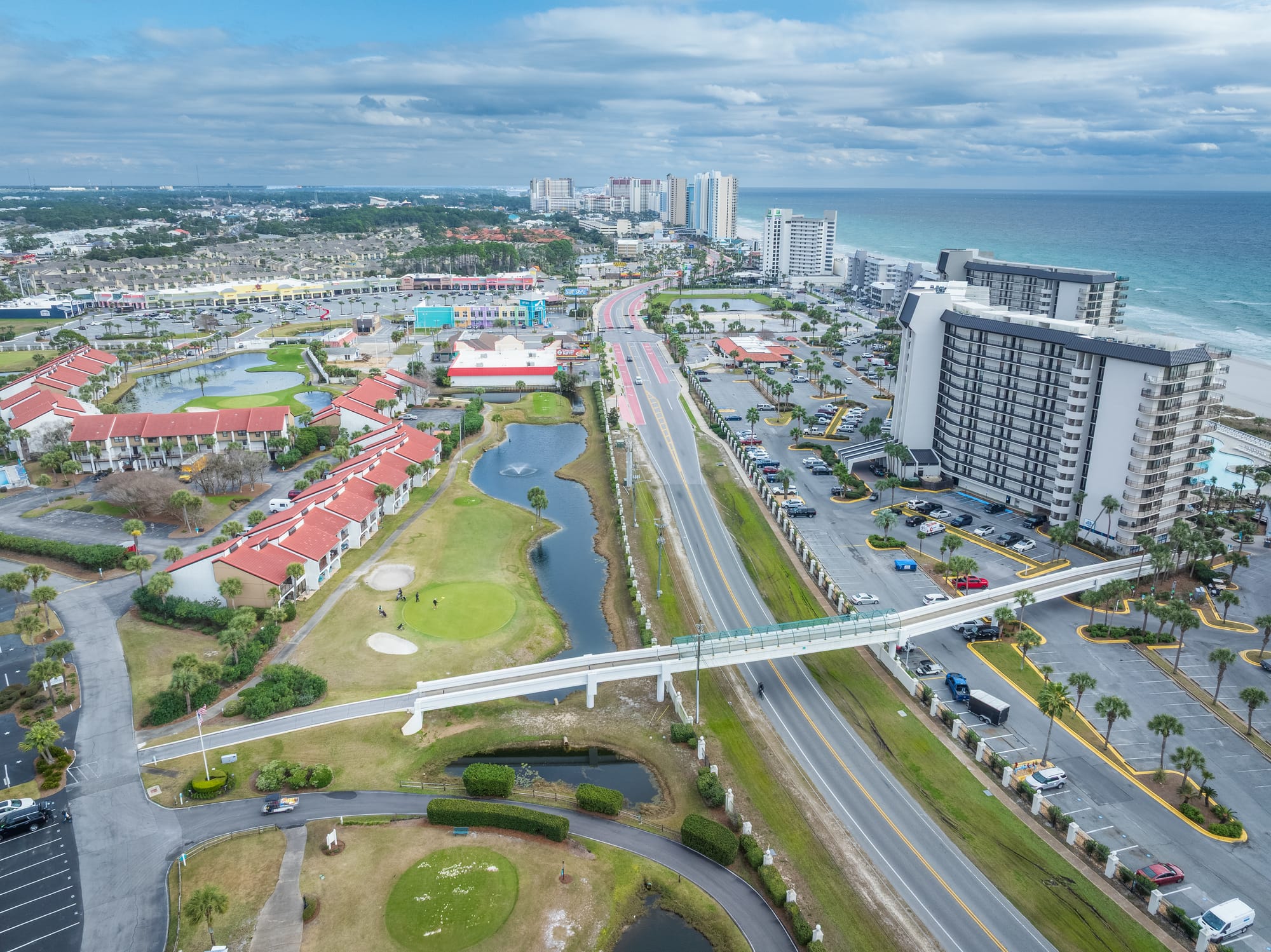 Aerial of the road, a pedestrian bridge, golf greens, and beachfront towers along the gulf.